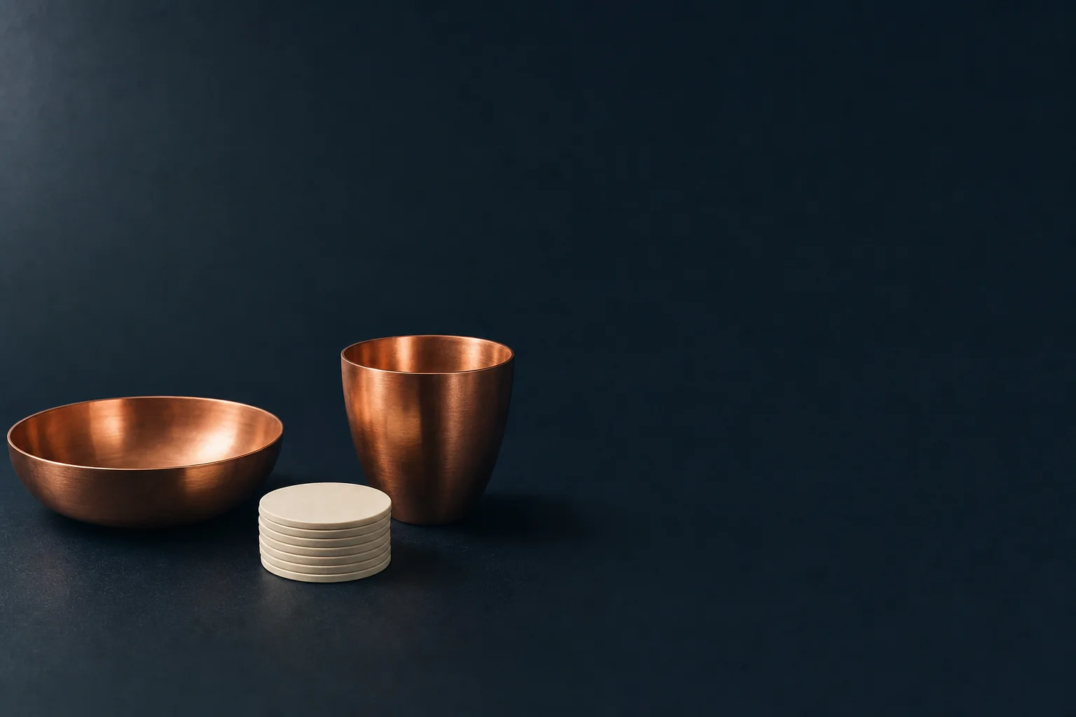 A shallow copper bowl and a deep copper cup on a navy desk with a short stack of blank ivory discs between them