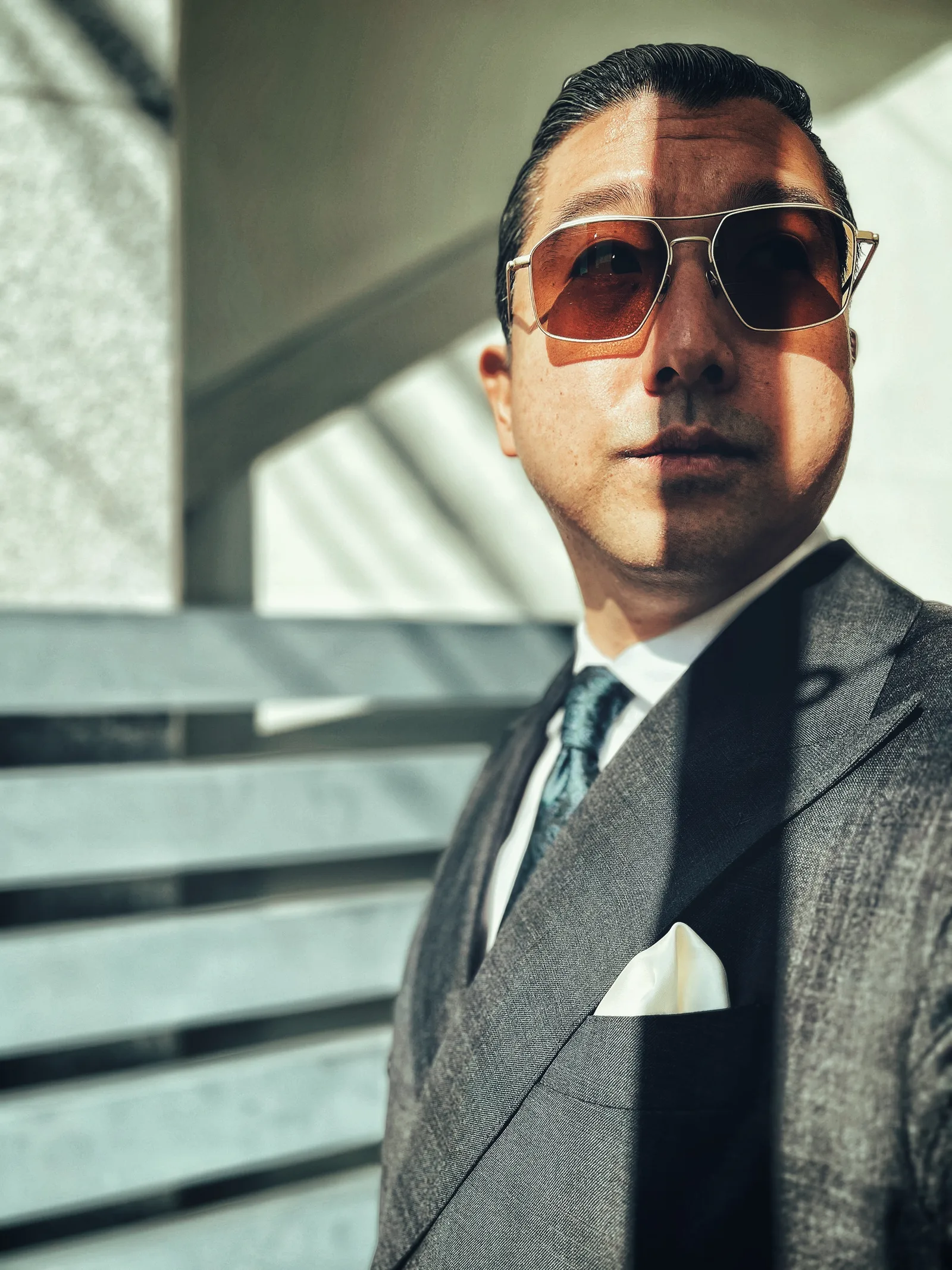 Joon K Lee in a grey suit and sunglasses, half his face in hard sunlight, half in shadow